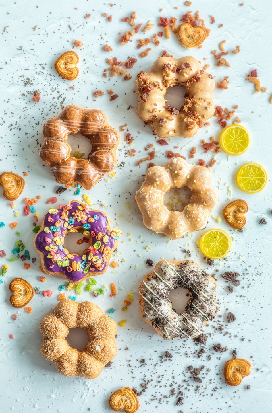 Six decorated mochi donuts arranged with scattered pastries and lemon slices.
